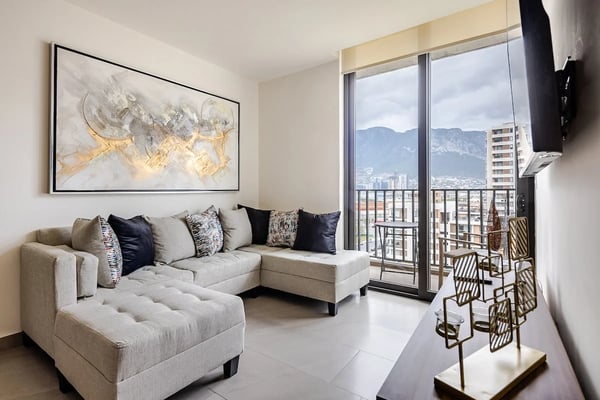 Sala moderna con sofá seccional beige, arte abstracto en la pared y ventanas de piso a techo con vistas a las montañas y la ciudad.